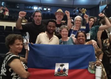 Un grupo diverso de personas en un evento en interiores portando la bandera haitiana y levantando los puños en un gesto de solidaridad.